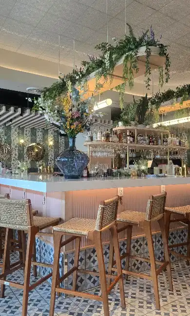 Suspended greenery shelf and tall floral vase above a restaurant bar with woven stools